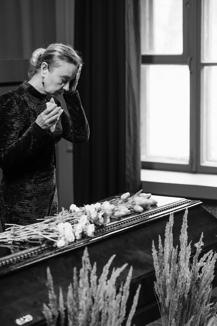 Woman mourning at a funeral, standing beside a coffin, holding flowers indoors.